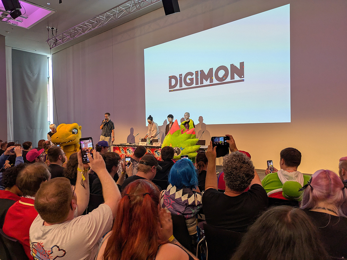 photo taken at the Toei Digimon panel. Agumon and the Chamelon type Digimon from Beatbreak are in front of the small stage while the crowd is cheering and taking photos