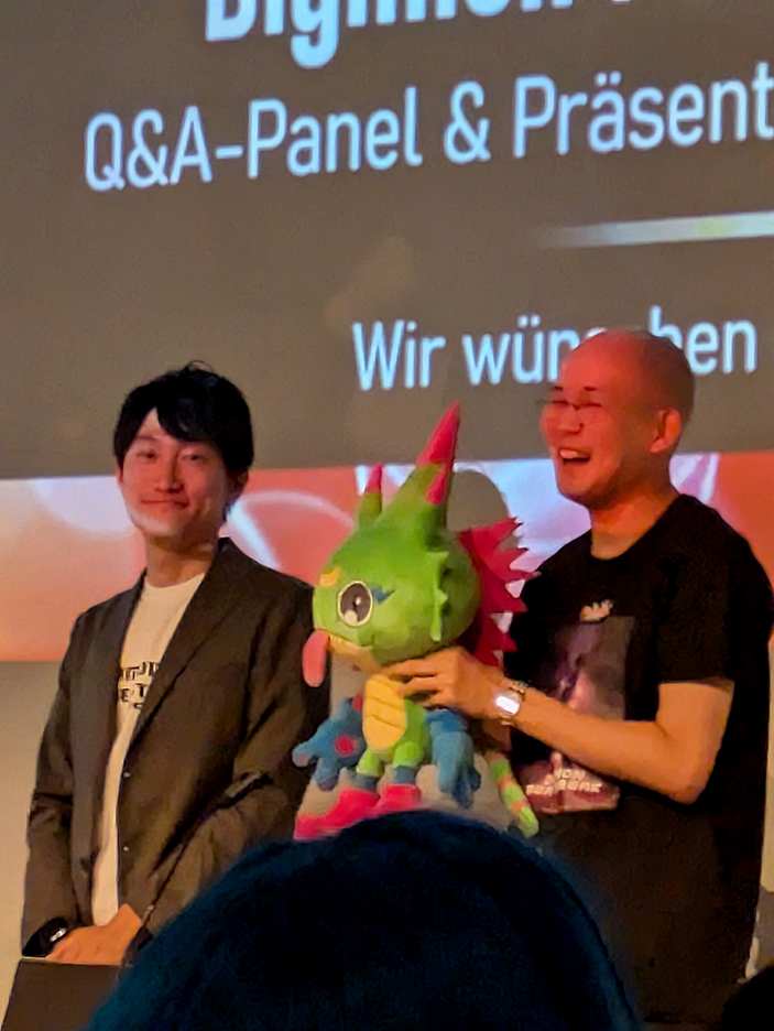 photo taken at the Toei Digimon panel: Nobutara Takada is smiling and holding a plush of Chamelon type Digimon from Beatbreak, Ryosuke Hara is standing beside him.